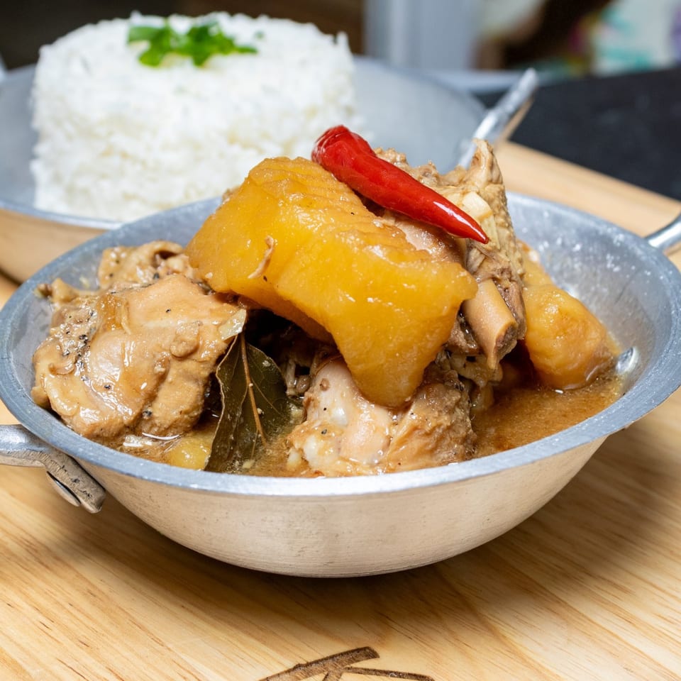 Filipino chicken adobo with potatoes and a red chili in a metal bowl, with rice in the background.