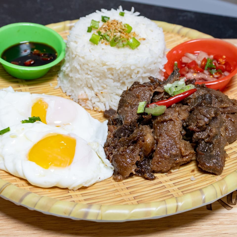 Filipino beef tapa with garlic rice, two fried eggs, and dipping sauces on a woven plate.