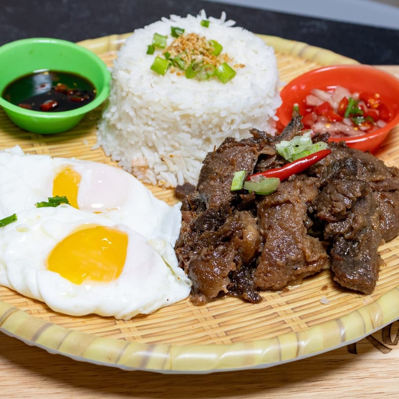 Filipino beef tapa with garlic rice, two fried eggs, and dipping sauces on a woven plate.