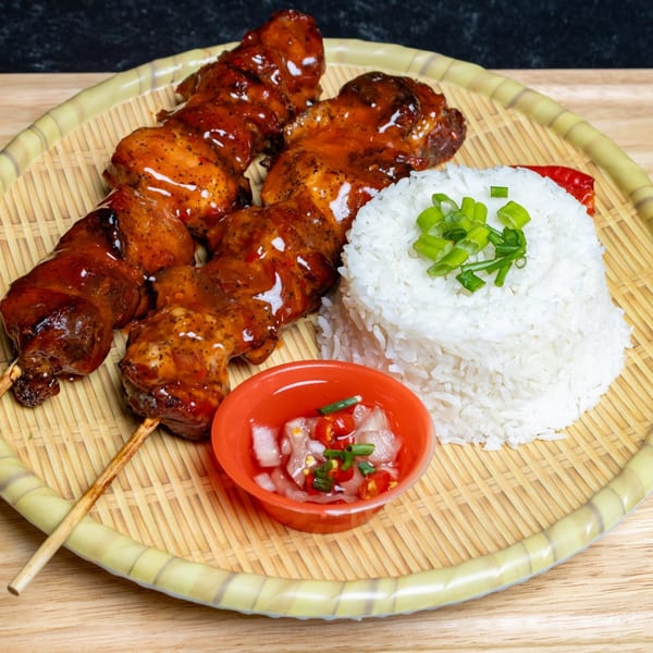 Two skewers of Filipino chicken barbecue with rice and a side of pickled onions and chilies.