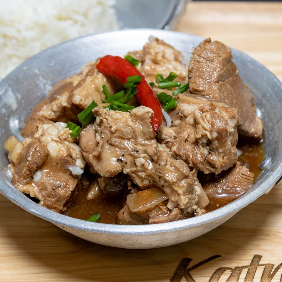 Filipino pork adobo with red chili and green onions in a metal bowl, with rice in the background.