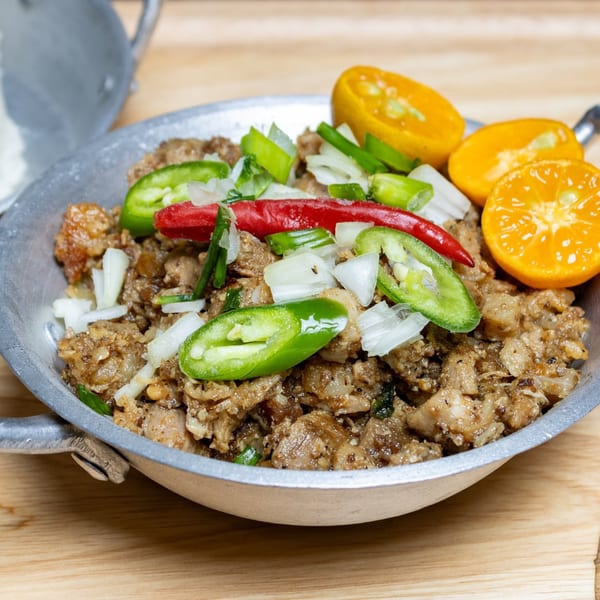 Filipino sisig in a metal skillet, garnished with red and green chilies, onions, and calamansi.
