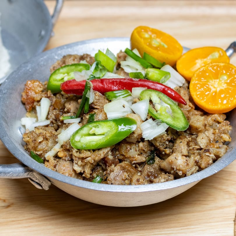 Filipino sisig in a metal skillet, garnished with red and green chilies, onions, and calamansi.