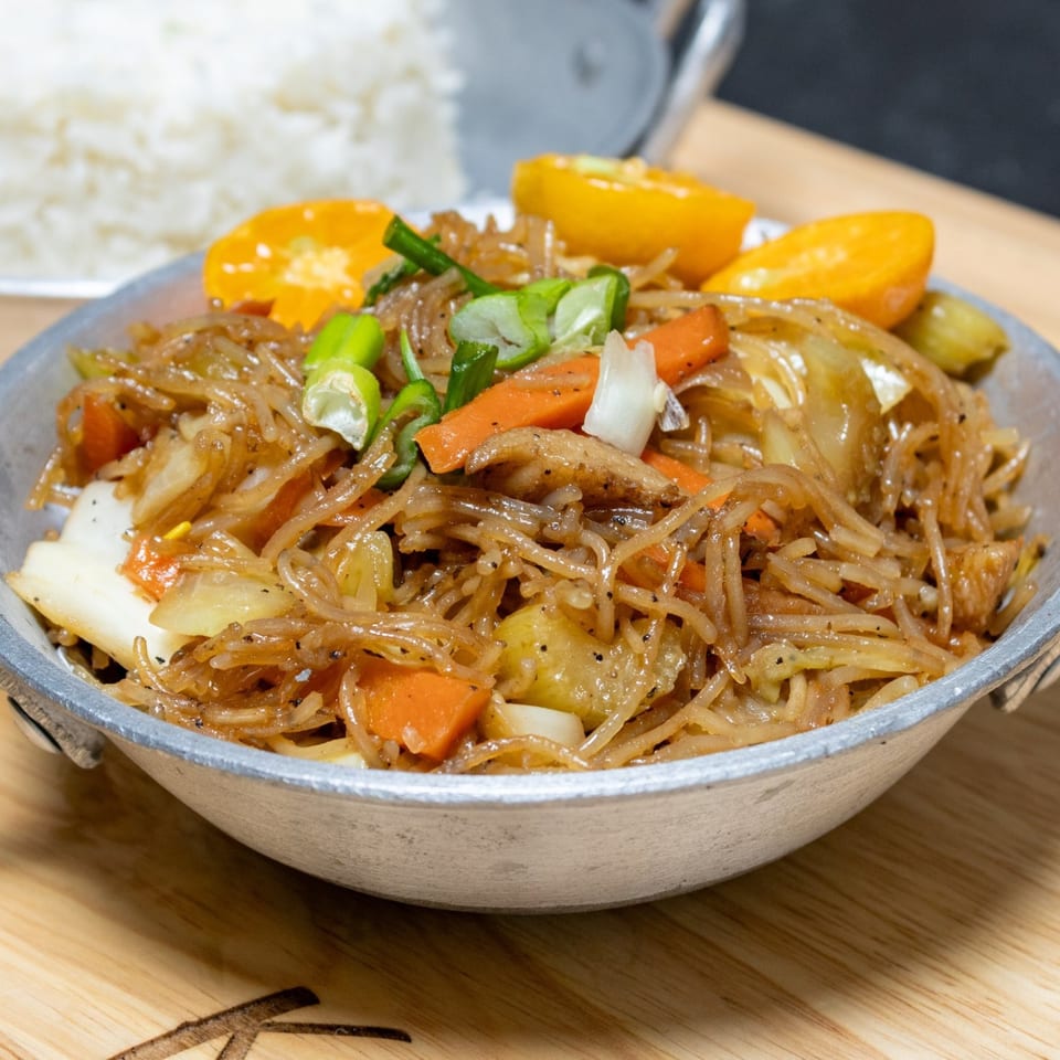 Filipino pancit bihon with vegetables and orange slices in a metal bowl, with rice in the background.