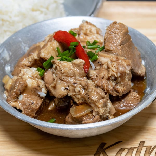 Filipino pork adobo in a metal bowl, garnished with red chili and green onions, with rice in the background.