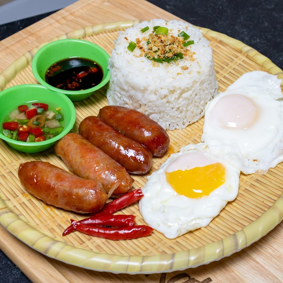 Filipino longganisa sausages, fried eggs, garlic rice, and dipping sauces on a woven plate.